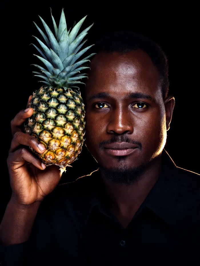 A Ugandan grower holding a ripe Smooth Cayenne pineapple beside his face, lit against black